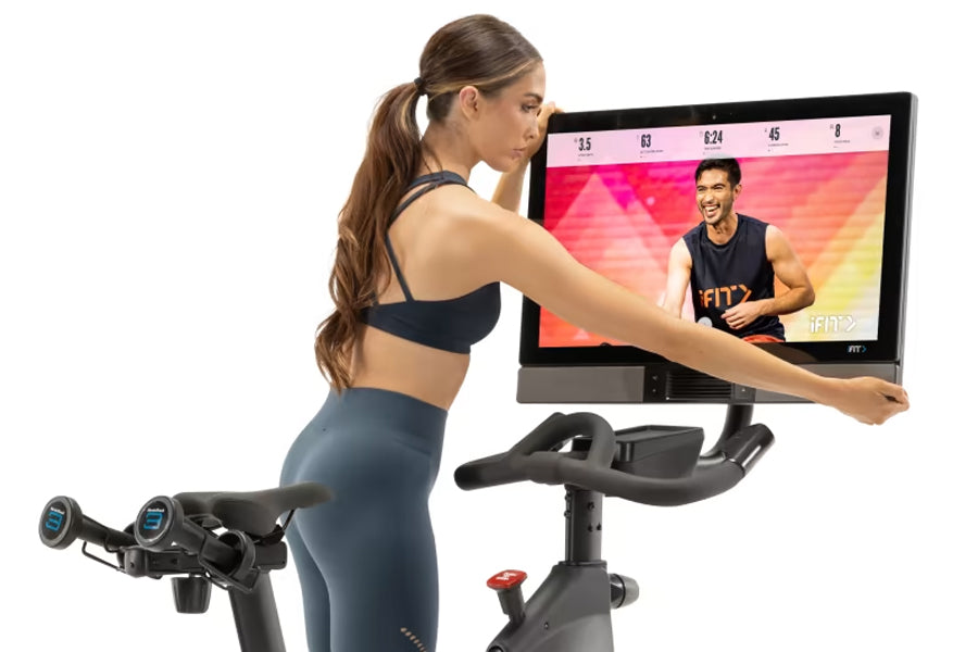 Woman in black sports bra and blue leggings using NordicTrack S27i stationary bike with attached screen displaying fitness cl