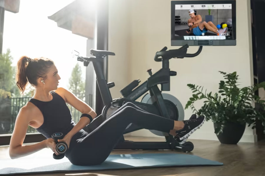 Woman in black tank top and leggings using stationary exercise bike with screen displaying fitness class, indoor home gym set