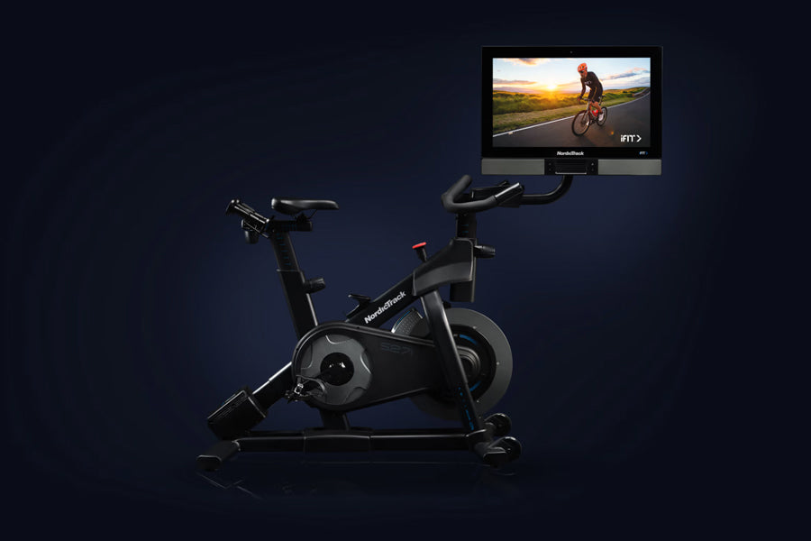 Black NordicTrack S27i stationary bike with integrated touchscreen display showing cycling workout video on dark background