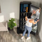 Black interactive home gym mirror with storage shelving, worn by model in white tank top and blue leggings in home setting
