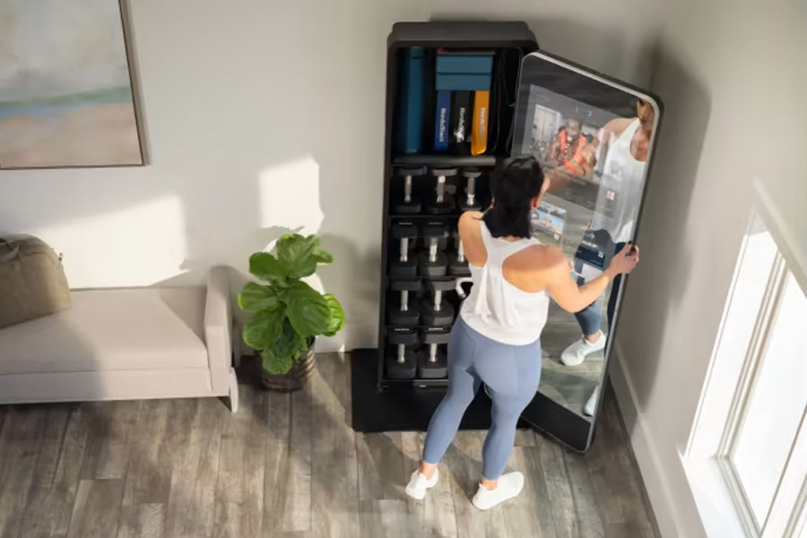 Black interactive home gym mirror with storage shelving, worn by model in white tank top and blue leggings in home setting