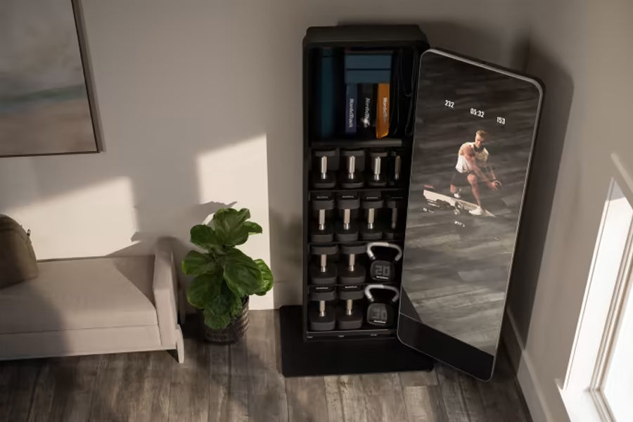 Black home gym mirror with integrated storage displaying fitness app on screen, flanked by potted plant and white furniture o