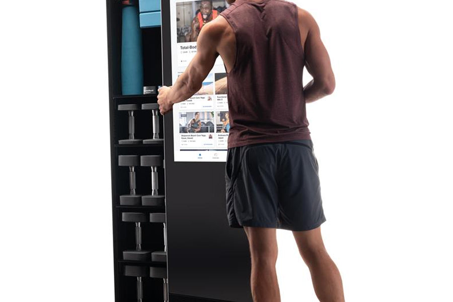 Man wearing burgundy sleeveless tank top and black shorts using NordicTrack Vault home gym mirror with dumbbell rack