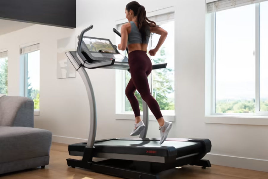 Woman running on NordicTrack X22i treadmill with digital display, wearing gray sports bra and burgundy leggings in bright hom