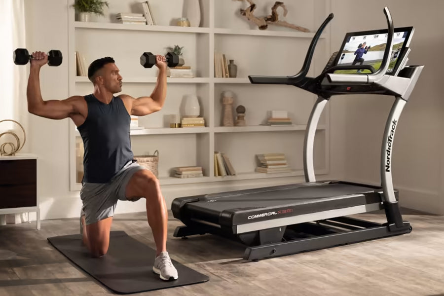 Man performing dumbbell shoulder press on black mat next to NordicTrack X32i treadmill with tablet display in home gym settin