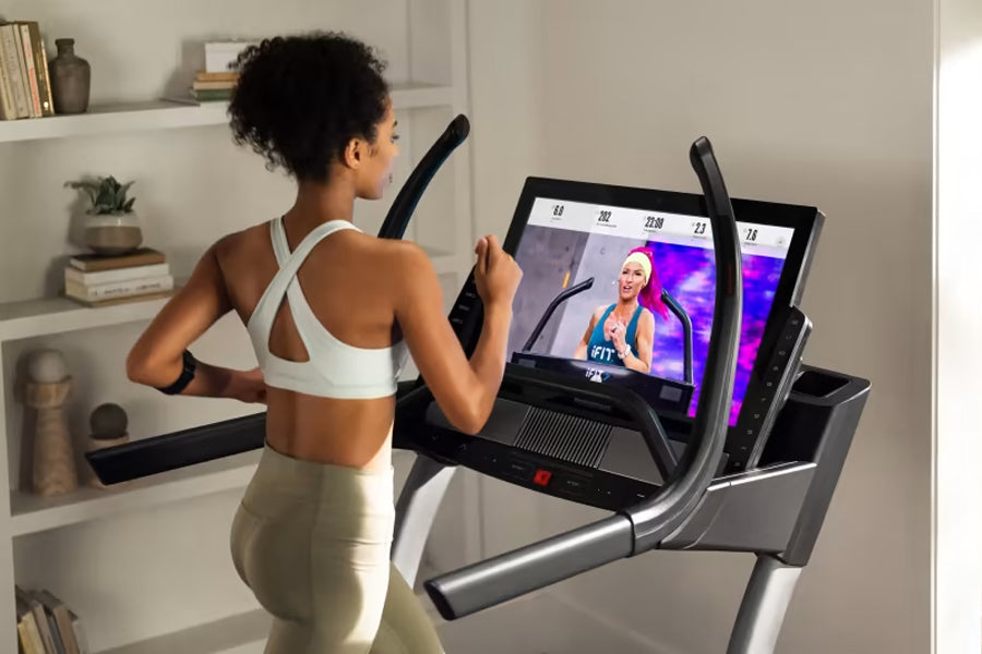 Woman using black and silver NordicTrack X32i treadmill with interactive screen display, wearing white sports bra and beige l