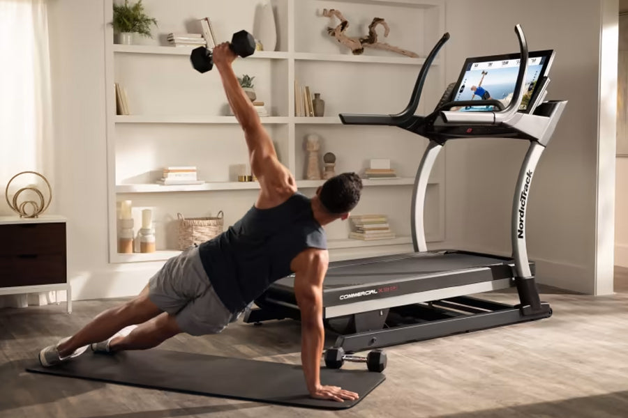 Man performing plank with dumbbell on black mat in front of NordicTrack X32i treadmill with touchscreen display in home gym