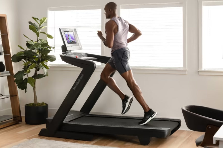 Man running on black NordicTrack treadmill with digital display in bright home gym with potted plant and windows