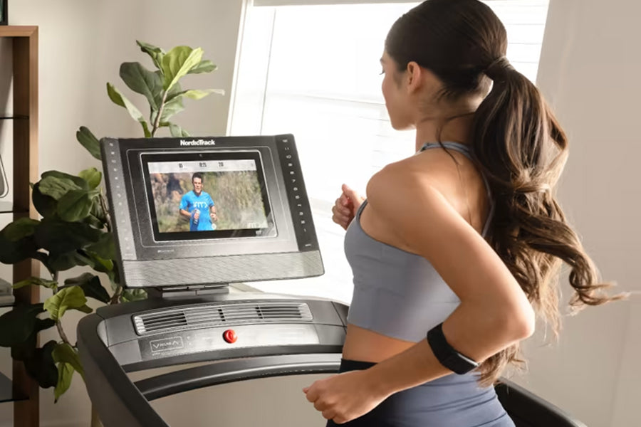 Woman in gray sports bra running on NordicTrack treadmill with digital display showing fitness video, potted plant visible in