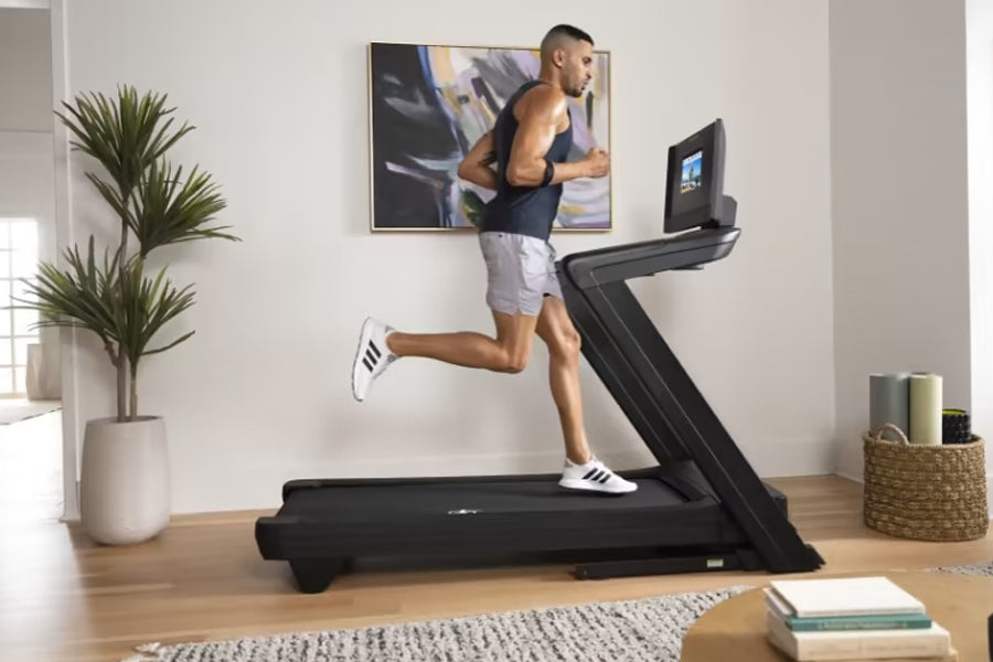 Man running on black NordicTrack treadmill with digital display in home gym setting, wearing white sneakers and gray shorts