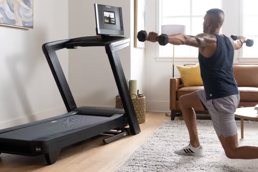 Man using black NordicTrack treadmill with digital display in home gym setting, holding dumbbells for workout