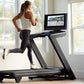 Woman running on black NordicTrack treadmill wearing white tank top and black leggings, with TV screen mounted above in brigh