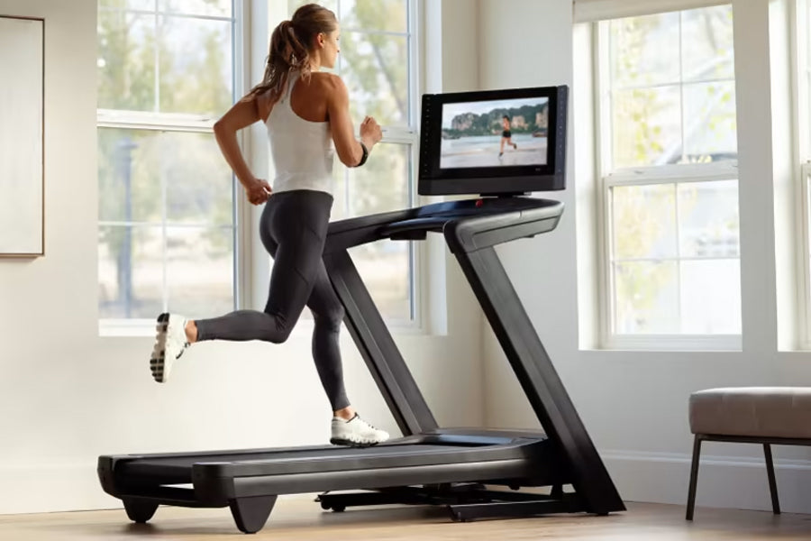 Woman running on black NordicTrack treadmill wearing white tank top and black leggings, with TV screen mounted above in brigh