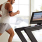 Man in white tank top and gray shorts running on black treadmill with touchscreen displaying beach scene, bright window backg