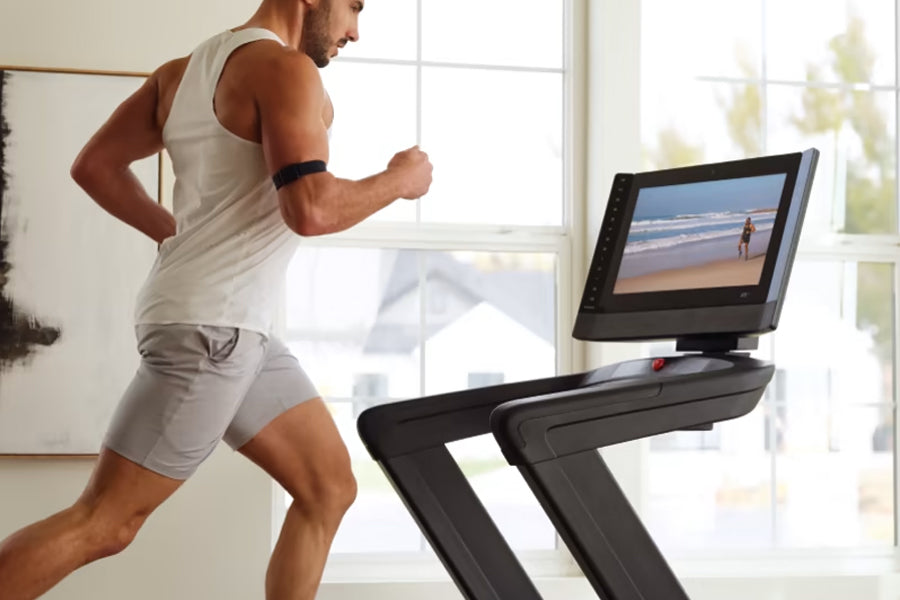 Man in white tank top and gray shorts running on black treadmill with touchscreen displaying beach scene, bright window backg