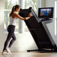 Woman using black NordicTrack treadmill with touchscreen display in bright home gym with large windows