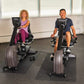 Two people using black and silver NuStep RB8MAX recumbent exercise bikes in a gym setting with exercise balls on rack behind 