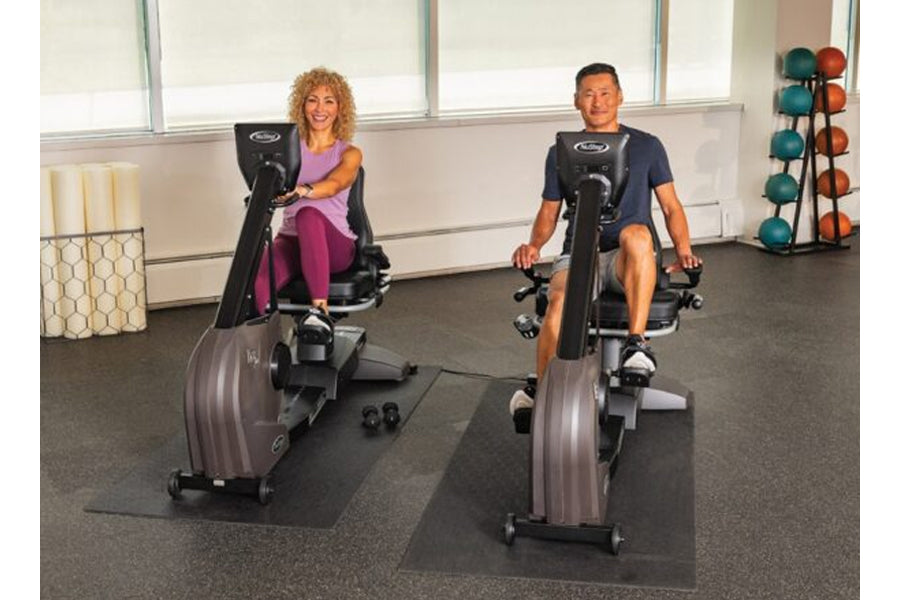 Two people using black and silver NuStep RB8MAX recumbent exercise bikes in a gym setting with exercise balls on rack behind 