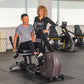 Man seated on black recumbent exercise bike with woman standing beside in gym setting with additional equipment visible