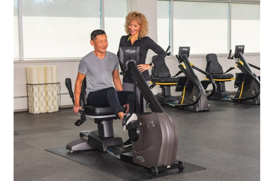Man seated on black recumbent exercise bike with woman standing beside in gym setting with additional equipment visible