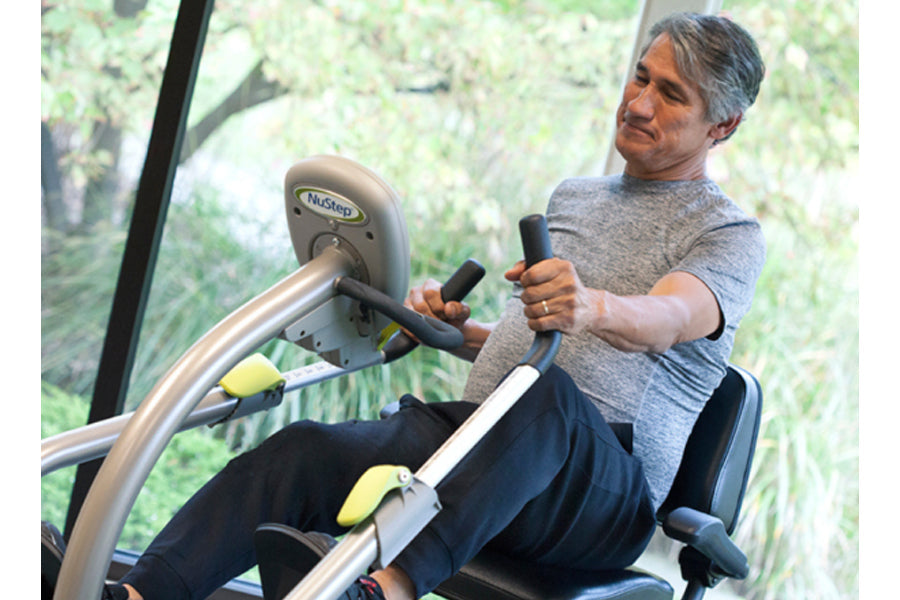 Man in gray shirt using NuStep T4r recumbent elliptical stepper with black seat and silver frame, near window with greenery