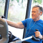 Man wearing blue t-shirt using NuStep recumbent elliptical stepper with earbuds, indoors near window