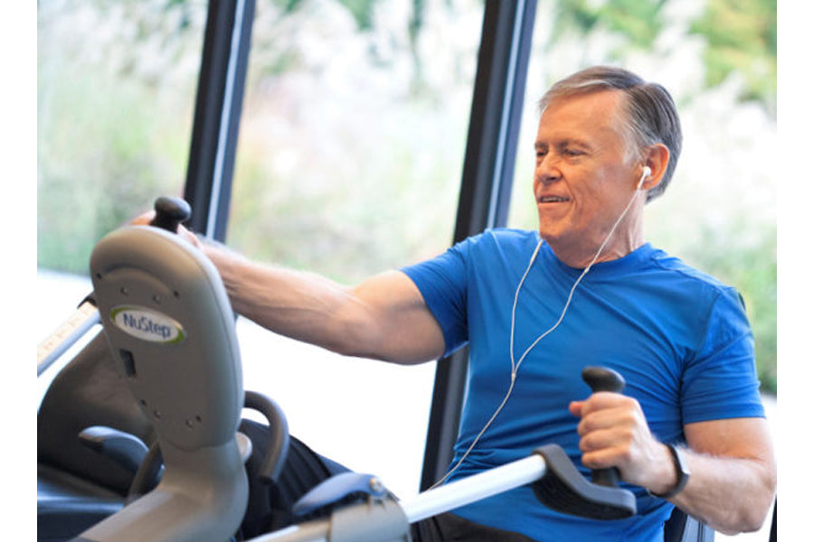 Man wearing blue t-shirt using NuStep recumbent elliptical stepper with earbuds, indoors near window