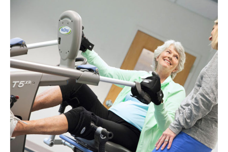Senior woman using NuStep T5XR recumbent elliptical stepper with green handles and black foot pedals, assisted by trainer in 