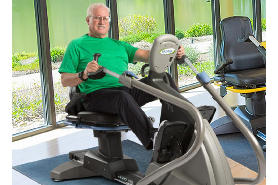 Older man in green shirt seated on gray NuStep recumbent elliptical stepper in bright fitness room with windows
