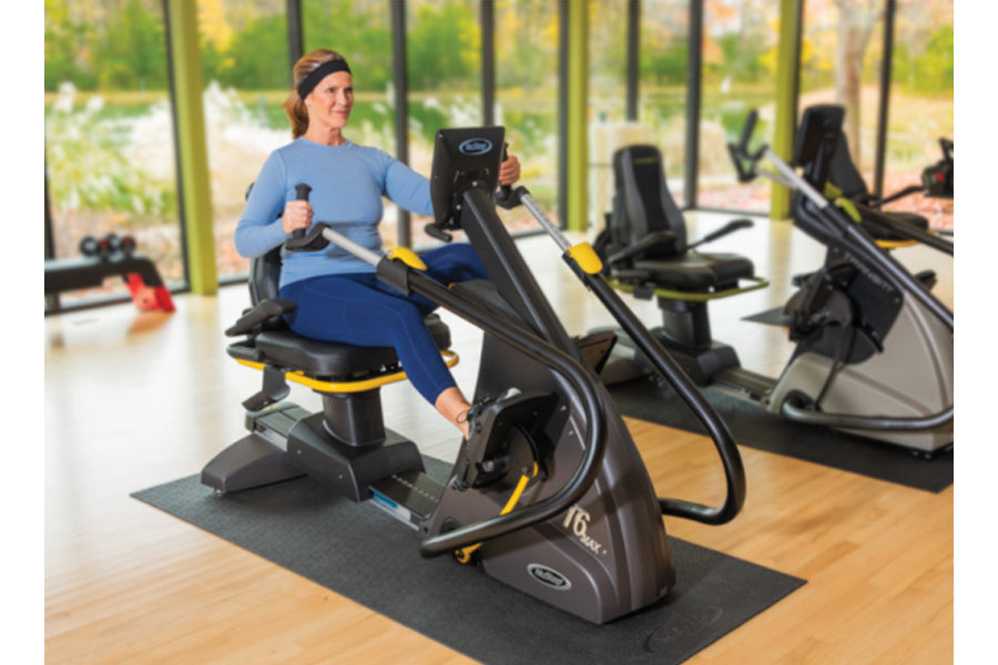 Woman in blue shirt and navy pants using black and yellow recumbent elliptical stepper in gym setting with windows