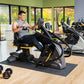 Man using black and yellow recumbent elliptical stepper in gym with large windows, dumbbells and additional equipment visible