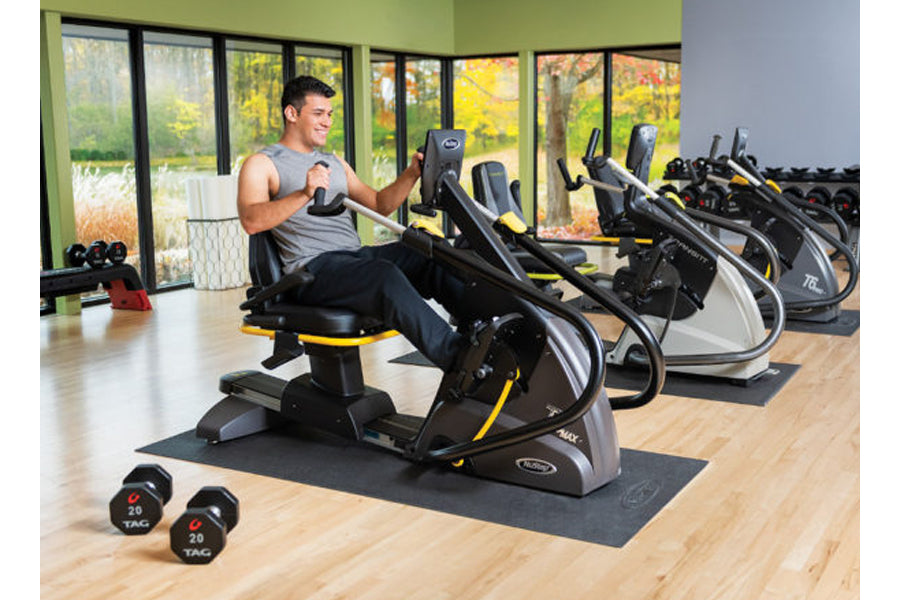 Man using black and yellow recumbent elliptical stepper in gym with large windows, dumbbells and additional equipment visible