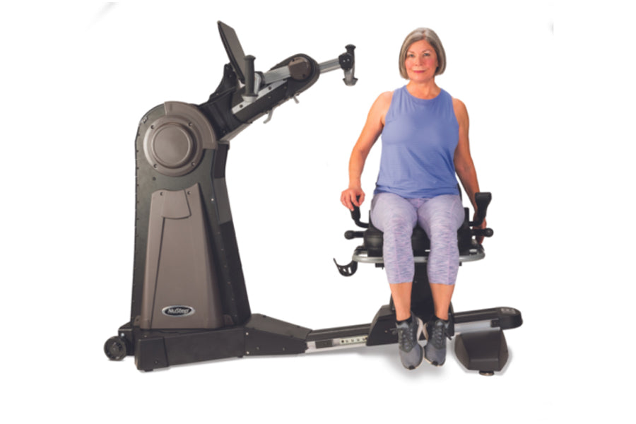 Woman seated on black and gray upper body ergometer machine with dual handles, wearing periwinkle sleeveless top and lavender