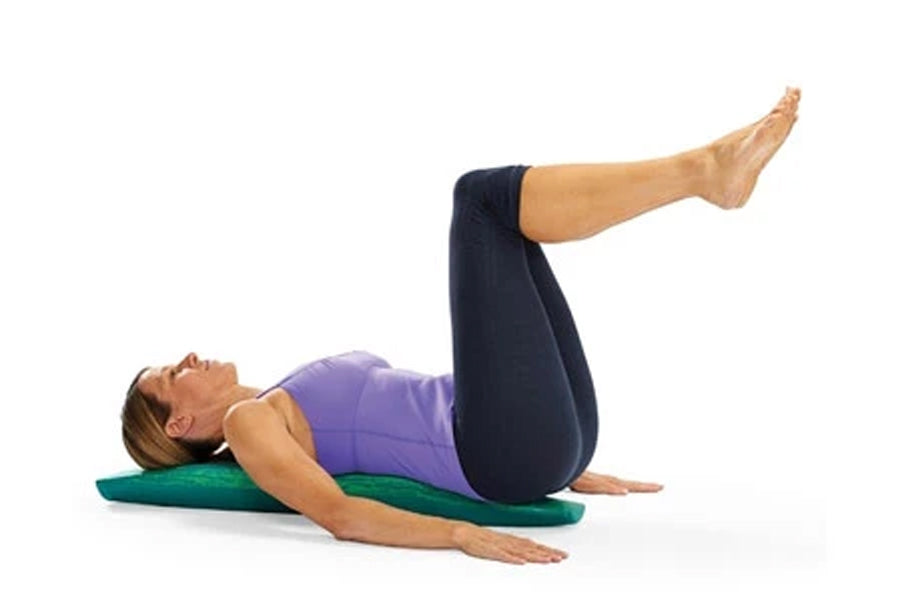 Woman lying on green foam roller wearing purple tank top and black leggings, demonstrating exercise position on white backgro