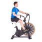 Man in blue t-shirt and black shorts riding white and black stationary air bike with orange fan wheel on white background