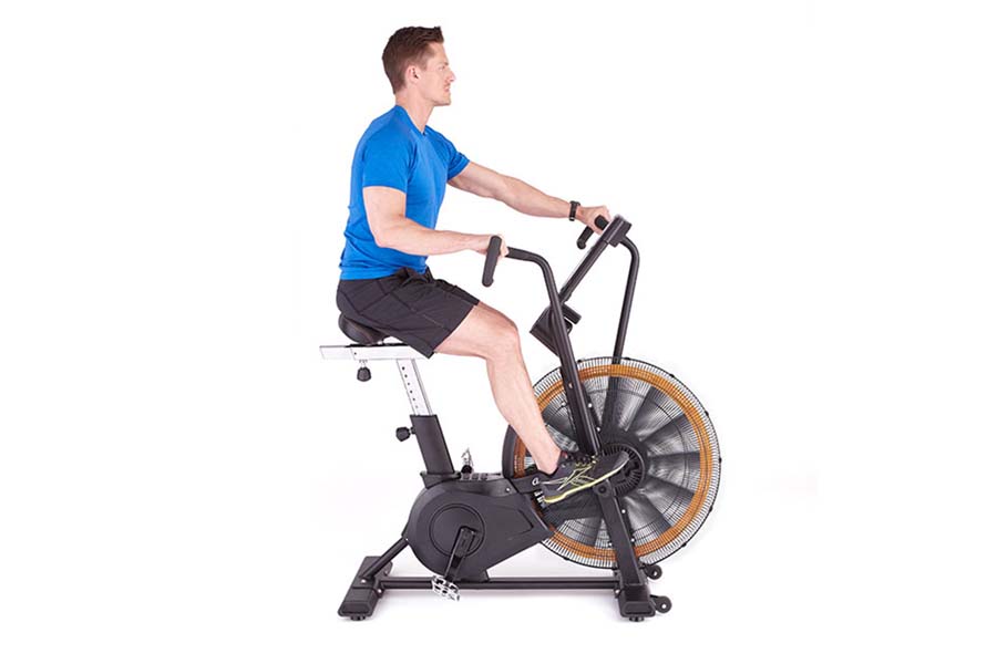 Man in blue t-shirt and black shorts riding white and black stationary air bike with orange fan wheel on white background
