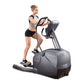 Woman wearing gray sports bra and black shorts using black and gray elliptical machine with touch screen console on white bac