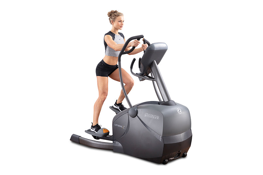 Woman wearing gray sports bra and black shorts using black and gray elliptical machine with touch screen console on white bac