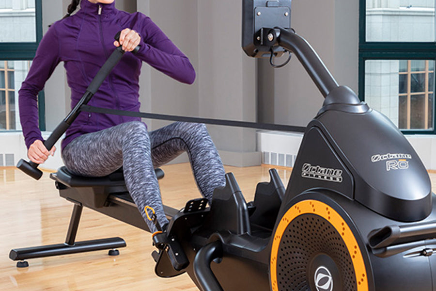Woman in purple jacket and gray leggings using black and orange Octane RO rowing machine indoors