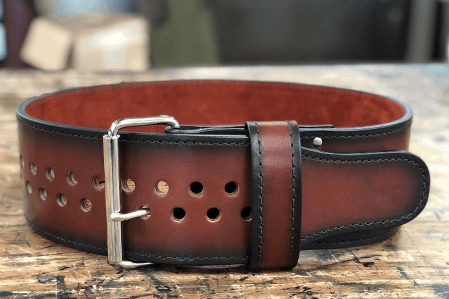 Brown leather belt with black edges and silver buckle, featuring double row of brass grommets, displayed on wooden surface