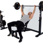 Man performing barbell bench press on black Powertec Olympic weight bench with gold-accented weight plates, dumbbell rack bes
