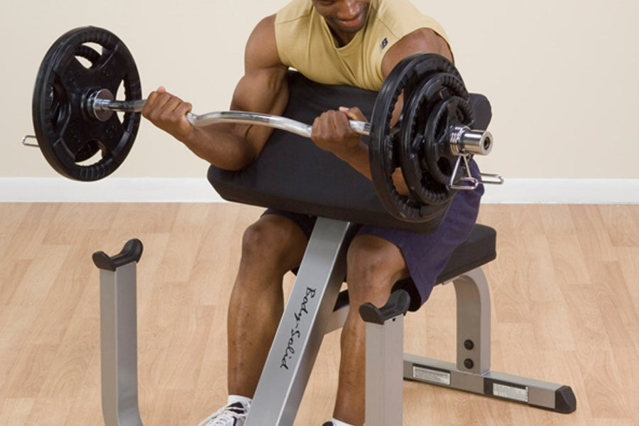 Man in yellow tank top performing bicep curl with barbell on gray preacher curl bench with black weight plates