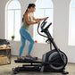 Woman using black ProForm Carbon EL elliptical machine indoors, wearing sage green tank top and blue leggings, holding smartp