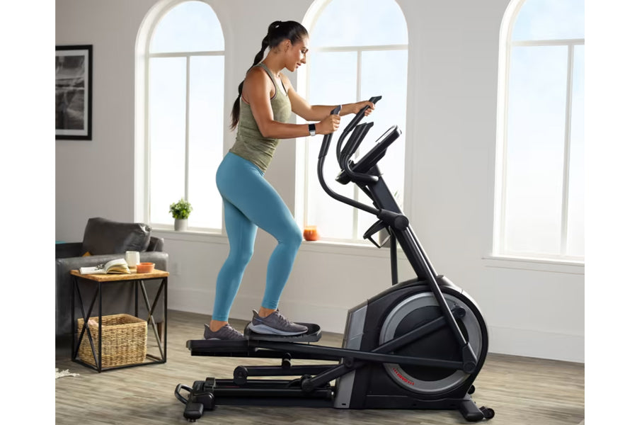 Woman using black ProForm Carbon EL elliptical machine indoors, wearing sage green tank top and blue leggings, holding smartp