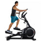 Man using black and gray ProForm Carbon EL elliptical machine, wearing teal shorts and black tank top on white background