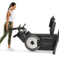Woman in white tank top and olive leggings using black ProForm Carbon HIIT H10 elliptical machine on white background