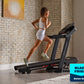 Woman running on black ProForm treadmill wearing white sports bra and shorts, in modern brick-walled home gym with plant