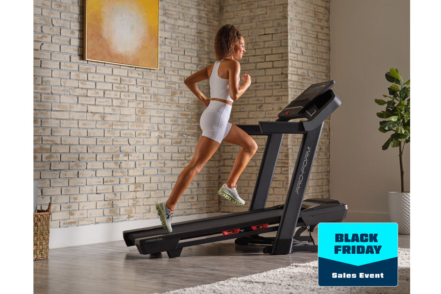 Woman running on black ProForm treadmill wearing white sports bra and shorts, in modern brick-walled home gym with plant