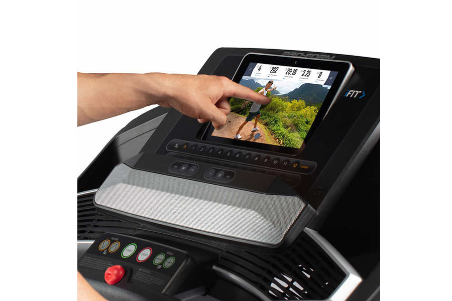 Hand touching touchscreen display on black ProForm treadmill showing outdoor running scene, with control panel and red emerge