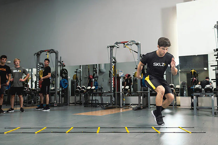 Athlete performing footwork drill with yellow and black agility ladder on gym floor, wearing SKLZ branded gear